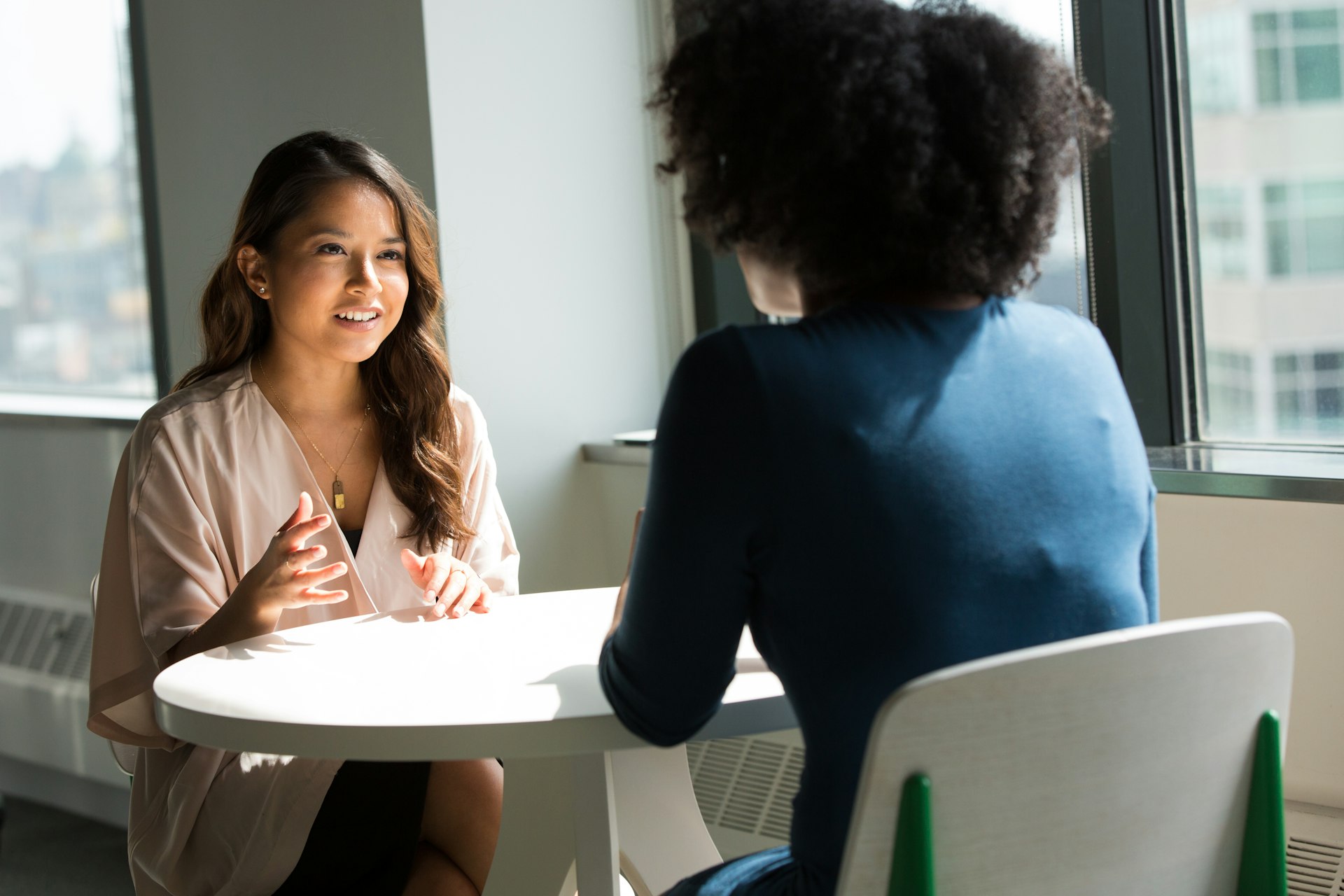 Deux femmes échangent autour d’une table près d’une grande fenêtre : une consultante IA écoute les besoins de sa cliente